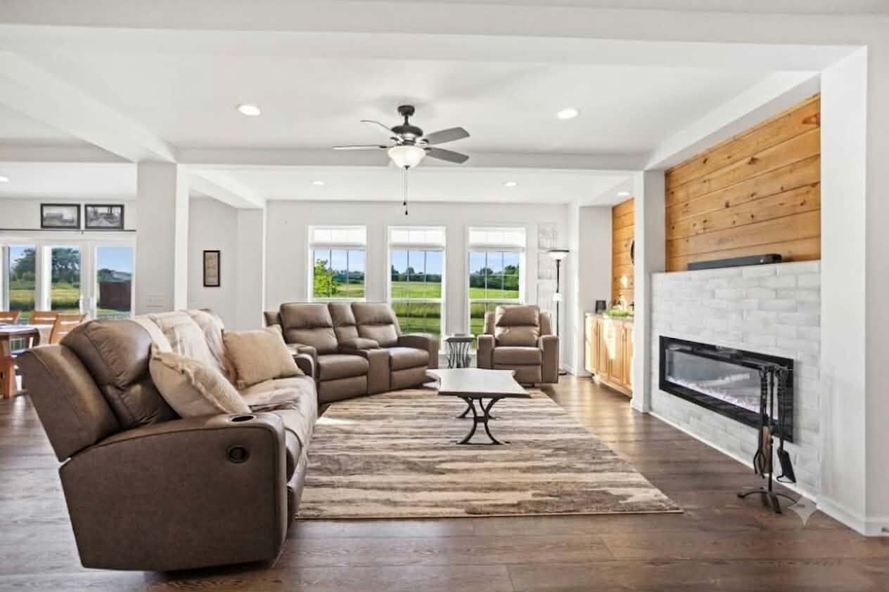 A spacious living room with a fireplace, a ceiling fan, and a large window offering a view of the outdoors.