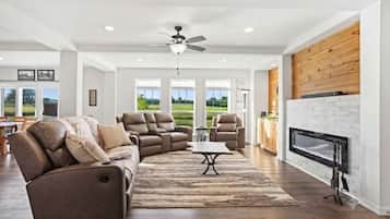 A spacious living room with a fireplace, a ceiling fan, and a large window offering a view of the outdoors.