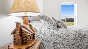 Ein Schlafzimmer mit einer lampenartigen Holzhaus-Form, einem Bett mit Bettdecke mit Baumzweig-Muster und einem Fenster mit Blick auf Grünflächen.