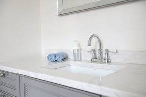 Ein Badezimmer mit einem weißen Marmorwaschtisch, einem silbernen Wasserhahn und einem Spiegel darüber. Auf dem Waschtisch liegen zwei zusammengerollte Handtücher und ein Seifenspender.