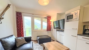 A compact living space with a sofa, ottoman, and a small wooden table. There is a kitchenette with a microwave and a TV mounted above it. The room has large windows with curtains and a view of greenery outside. - Justine (Top 9) by Interhome (Kaprun)
