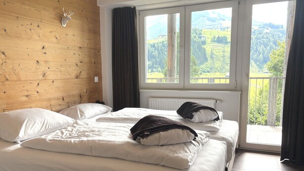 A bedroom with a wooden headboard, a large bed with white bedding, and a view of a green landscape through a window.