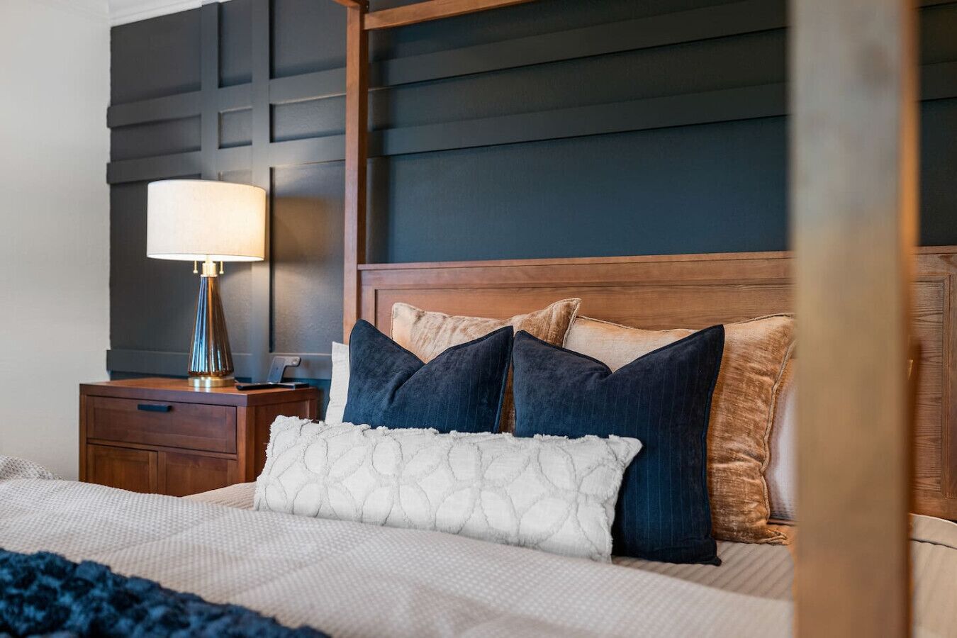 Une chambre d’hôtel avec une tête de lit en bois, une couvre-lit blanche à motifs et une table de chevet avec une lampe.