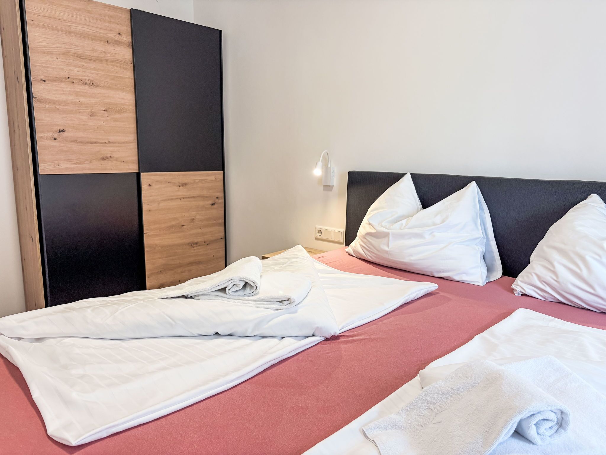 Ein ordentlich bezogenes Bett mit weißen Leinen, einer rosa Tagesdecke und einem Holzschrank mit einer schwarzen Furniereinlage.