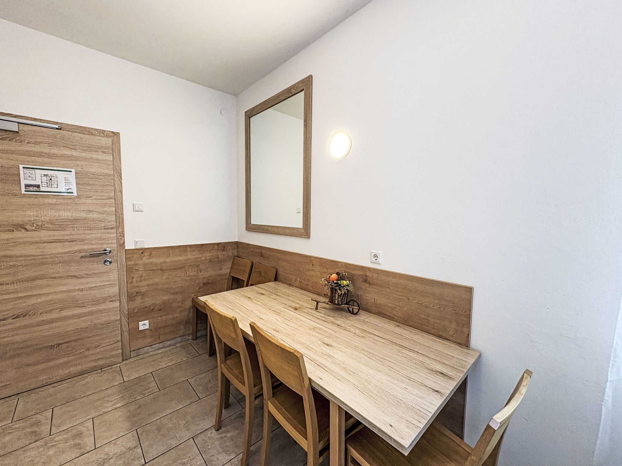 Une petite pièce, de taille modeste, avec une table et des chaises en bois, un miroir au mur et une porte dotée d’une poignée.