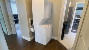 Une petite buanderie avec une machine à laver et un sèche-linge superposés, un plancher en bois et un coin repas visible à travers une porte ouverte.