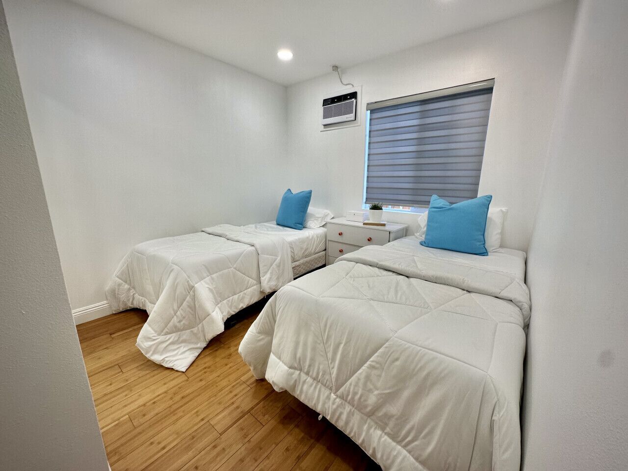 A bedroom with two beds, a window with blinds, a bedside table, and a wall-mounted air conditioner.
