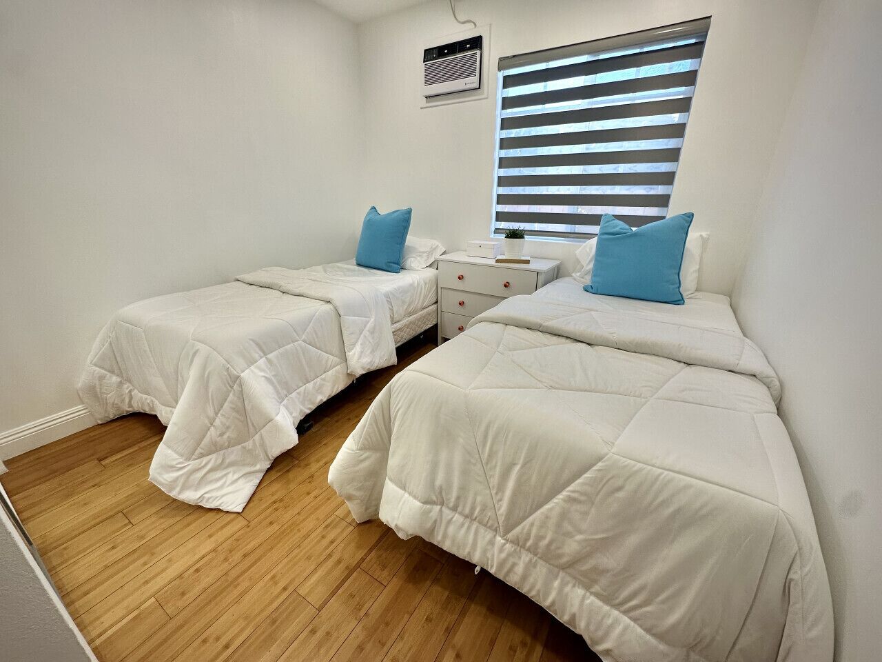 A bedroom with two beds, a small bedside table, a nightstand, and a window with blinds.
