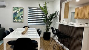 A modern, well-lit room with a white table and chairs, a large potted plant, a striped wall, and a kitchen area with wooden cabinets.