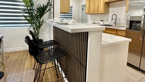 A modern kitchen with wooden cabinets, a stainless steel refrigerator, and a small island with a sink. There are two black chairs and a potted plant near the window.