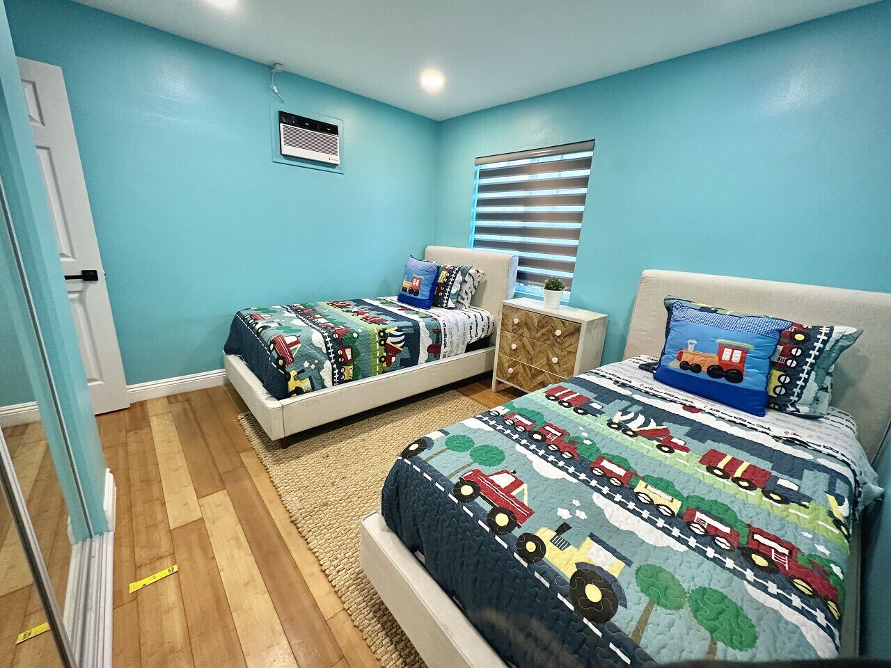 Un dormitorio con dos camas, cada una con una colcha de diseño alegre con motivos de tren, una ventana con persianas y una pequeña mesita de noche con lámpara.