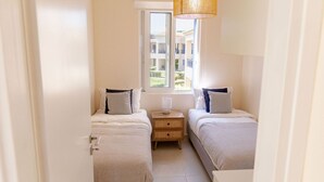 Un dormitorio pequeño y luminoso con dos camas individuales, una mesita de noche con lámpara, y una ventana que ofrece vistas a edificios.