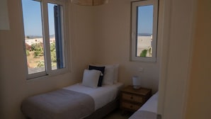 A small, single-bed room with a window, a bedside table, and a lamp.