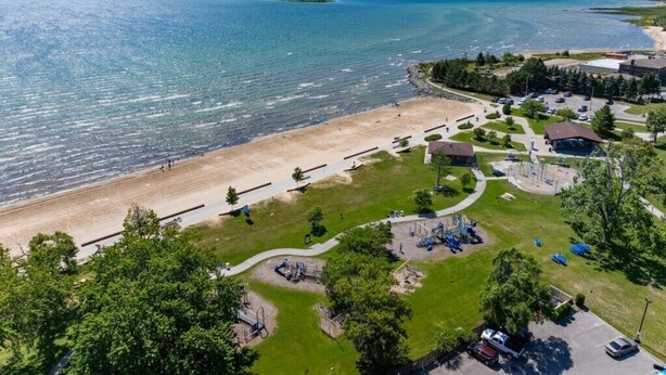 An aerial view of a coastal area with a beach, parking lot, playground, and buildings. - Lake Views | King Bed | Fenced (Alpena)