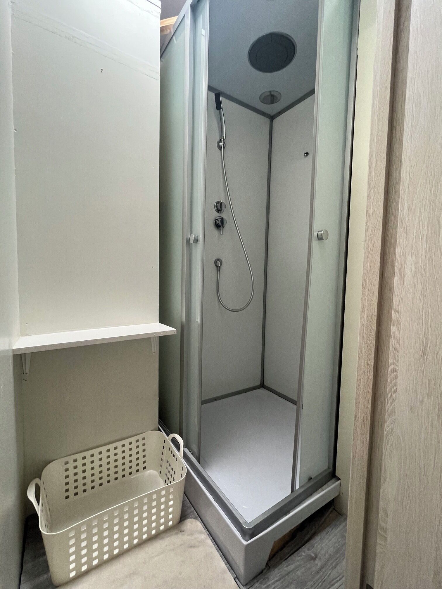 A small, enclosed shower area with a glass door, a metal showerhead, and a white plastic basket.