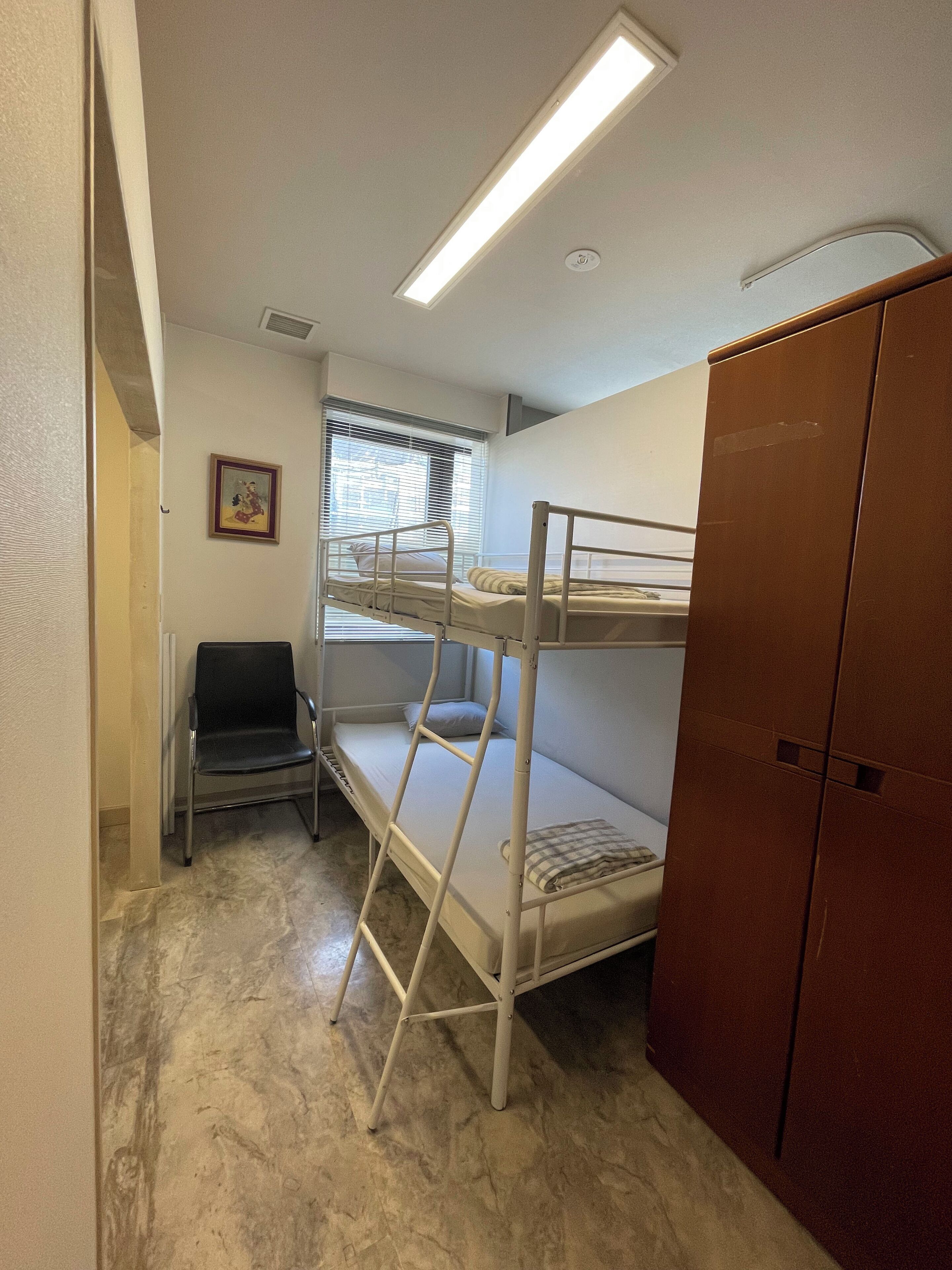 Quarto com beliche, guarda-roupa de madeira, uma cadeira e janela com persianas.