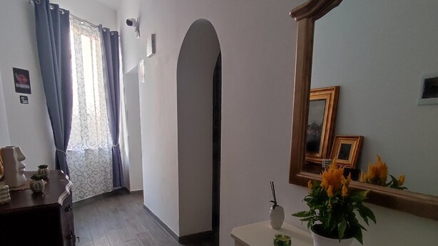 Um quarto com piso de madeira, uma toucador branca com espelho, uma mesa pequena com um vaso de flores e uma porta que dá para outro cômodo.