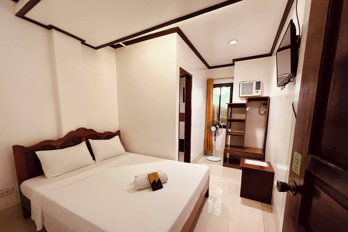 Quarto de hotel com cama, mesa de cabeceira, escrivaninha, cadeira, guarda-roupa e porta.