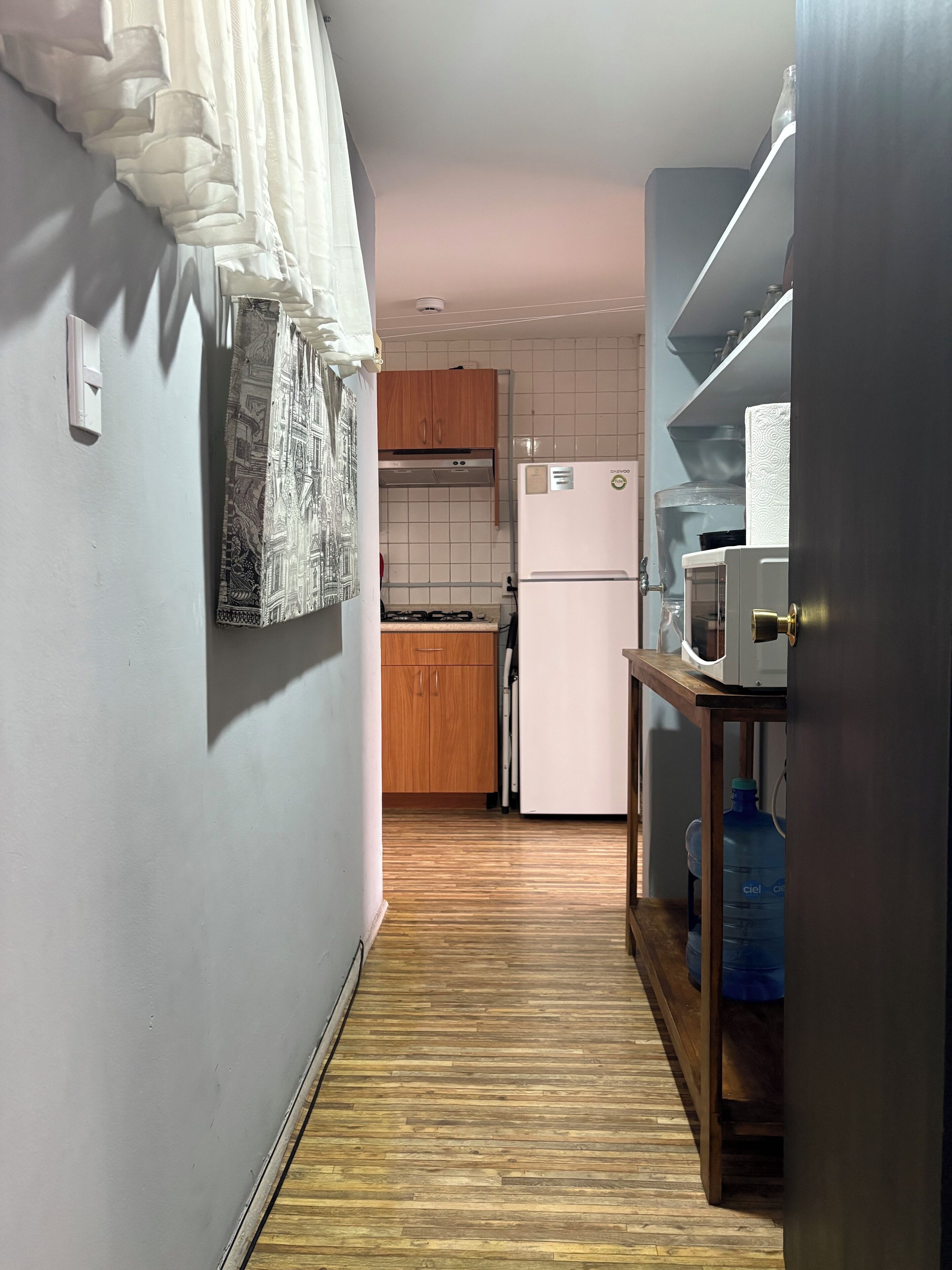 Una cocina estrecha con armarios de madera, un refrigerador blanco y una mesa de madera con dispensador de agua.