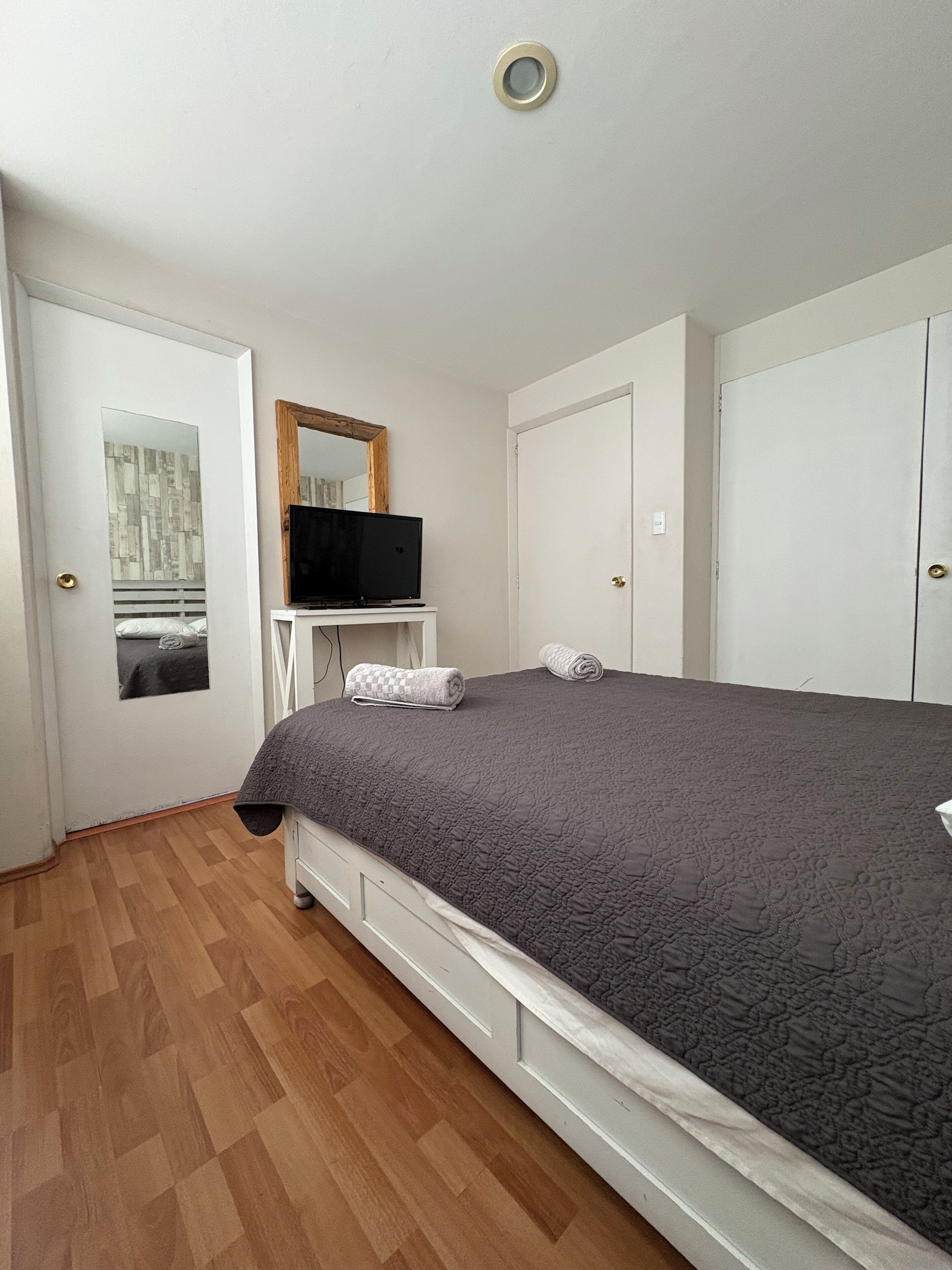 A bedroom with a bed, a TV on a stand, a wardrobe, and a mirror on the wall.