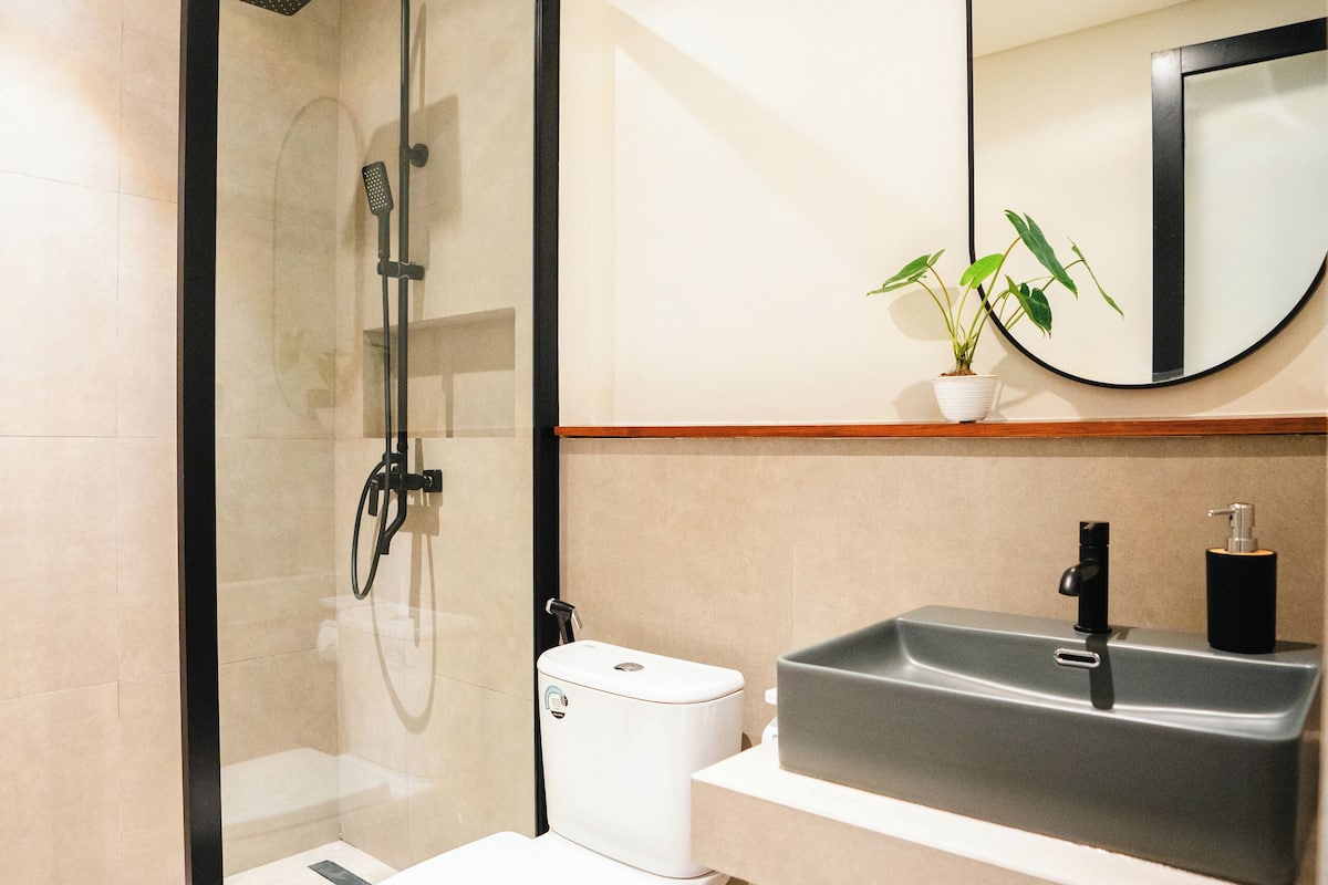 A modern bathroom with a glass-enclosed shower, a white toilet, a grey sink with a mirror above it, and a shelf with towels.