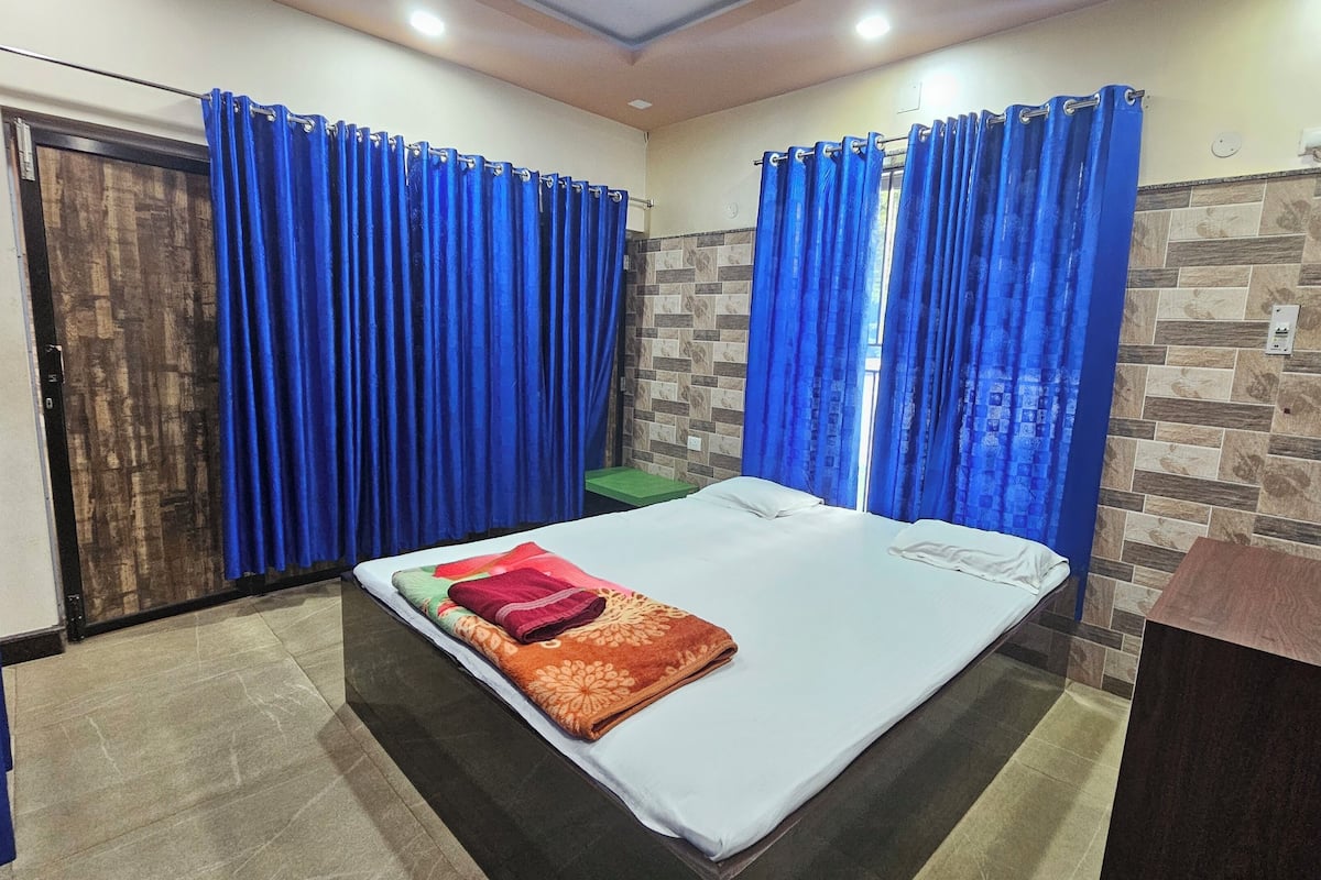 Un dormitorio con cama, cortinas azules, un taburete verde pequeño y una botella de agua sobre una mesa de madera.