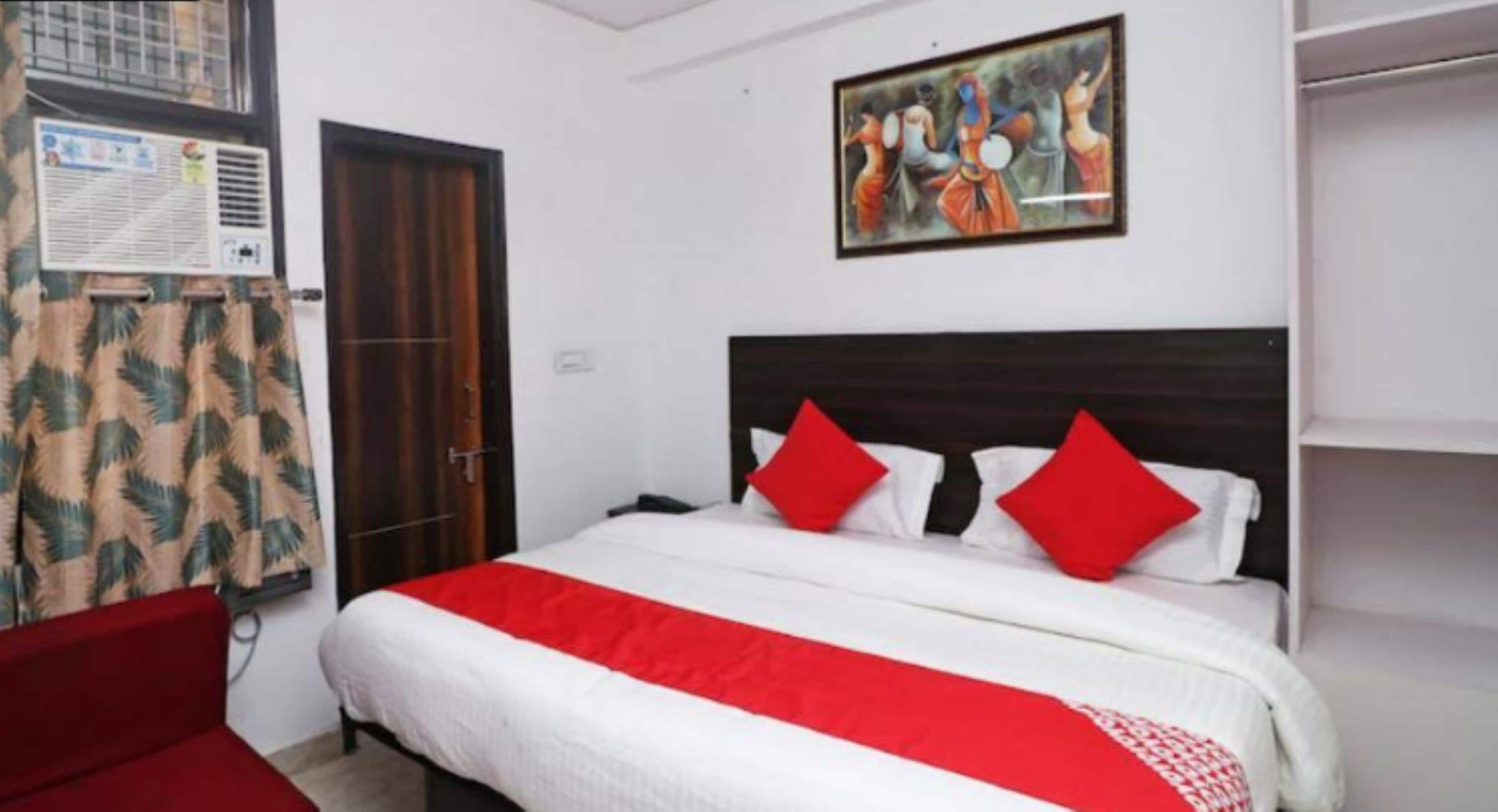 A hotel room with a bed, a red and white bedspread, a red sofa, a wooden door, a window with a curtain, and a painting on the wall.