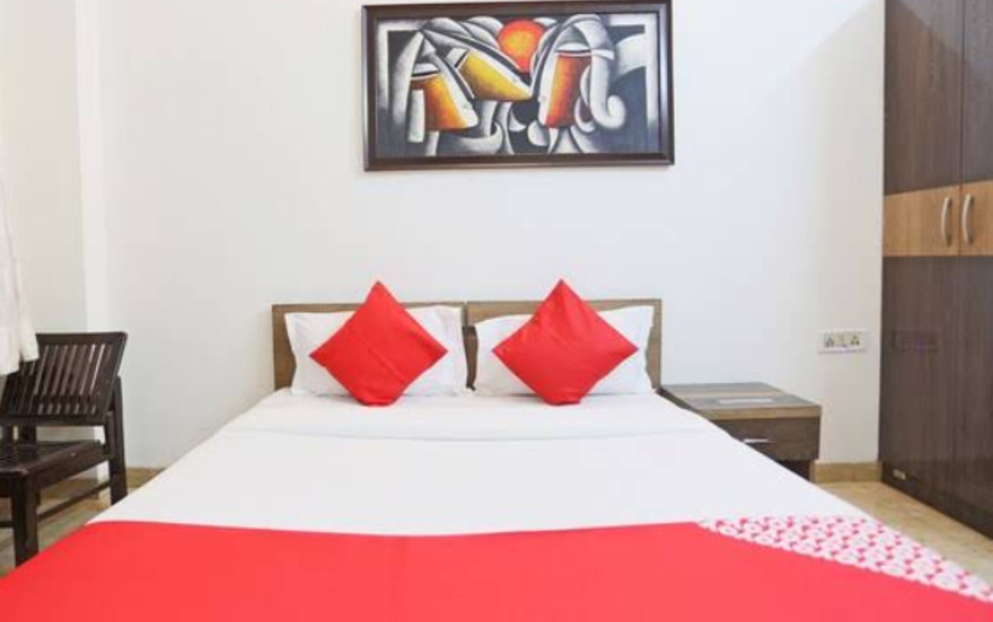 Una habitación de hotel con una cama, dos almohadas rojas, una colcha blanca, una manta roja, una silla de madera, una mesita de noche, un armario y un cuadro en la pared.