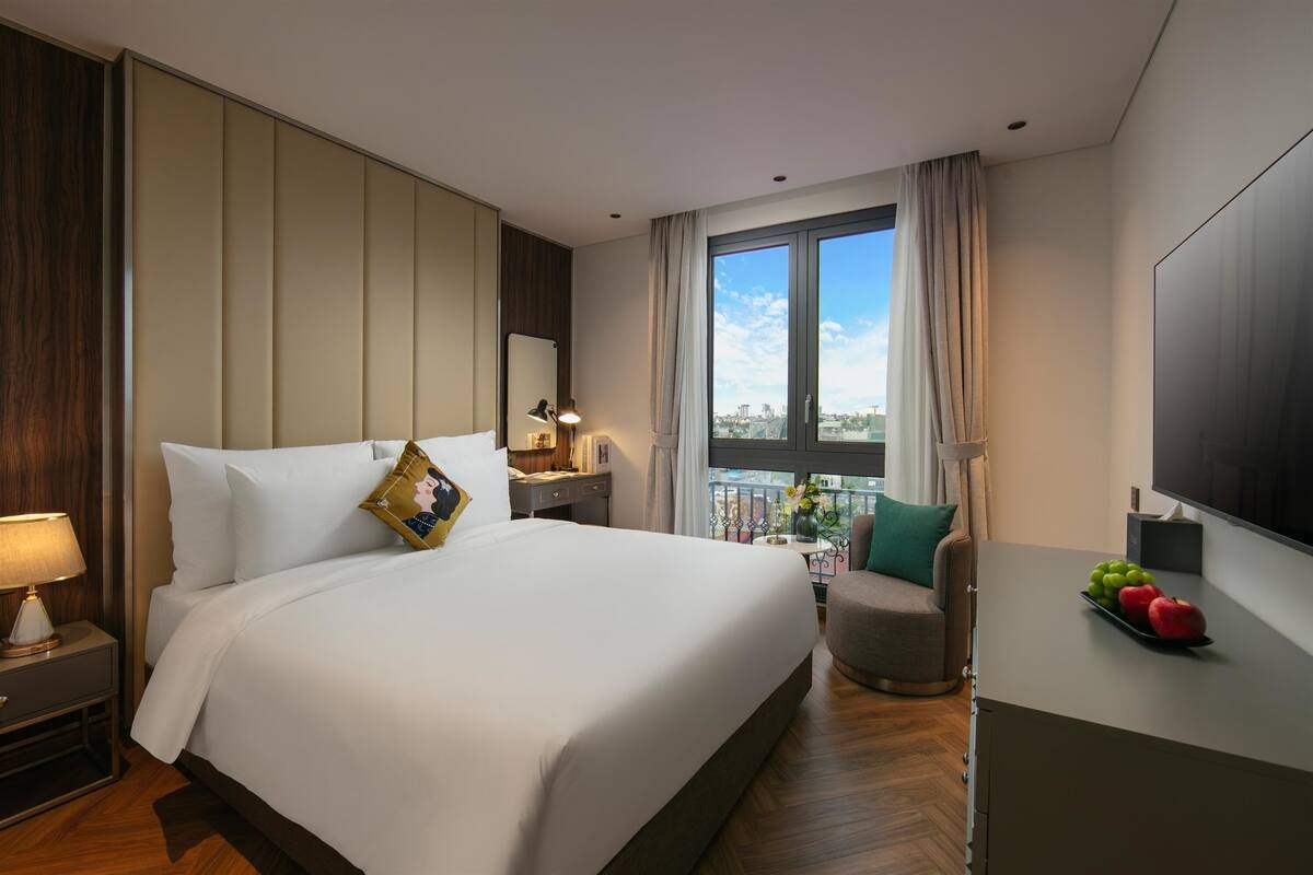 A hotel room with a large bed, a desk with a lamp, a view of the city through a window, and a TV on the wall.
