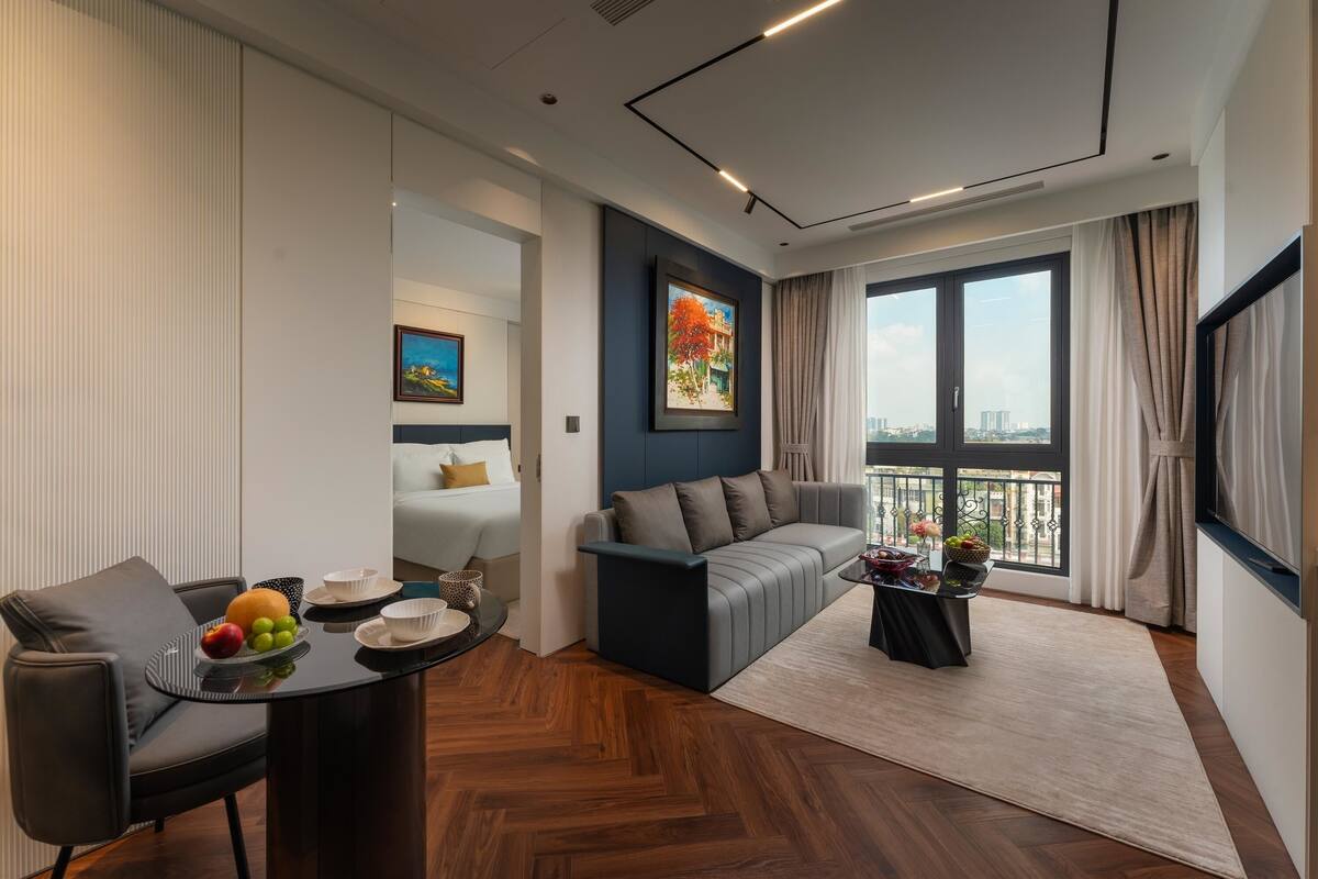 A modern hotel room with a sofa, a dining table with fruit, a view of the city, and a large window.