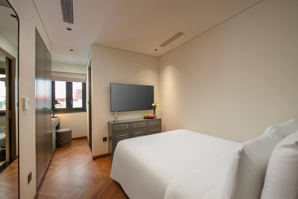 A hotel room with a bed, a TV mounted on the wall, a small table with a vase of flowers, and a window with a view of buildings.