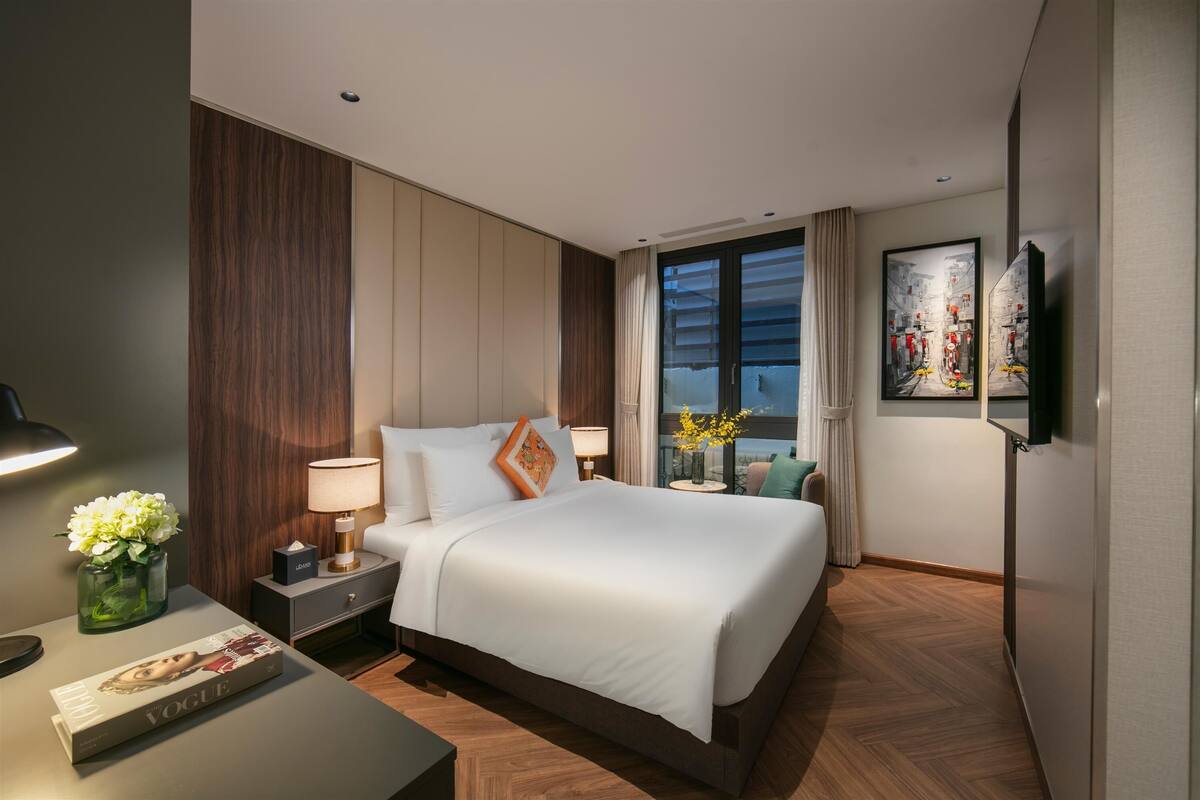 A modern hotel room with a large bed, a bedside table with a lamp, a vase with flowers, and a magazine on the nightstand.
