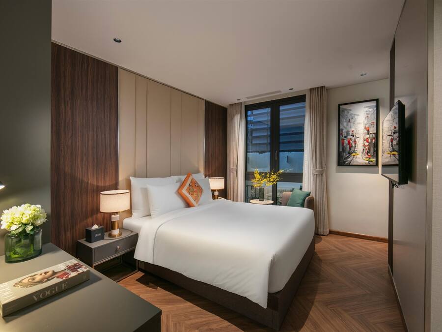 A modern hotel room with a large bed, a bedside table with a lamp, a vase with flowers, and a magazine on the nightstand.