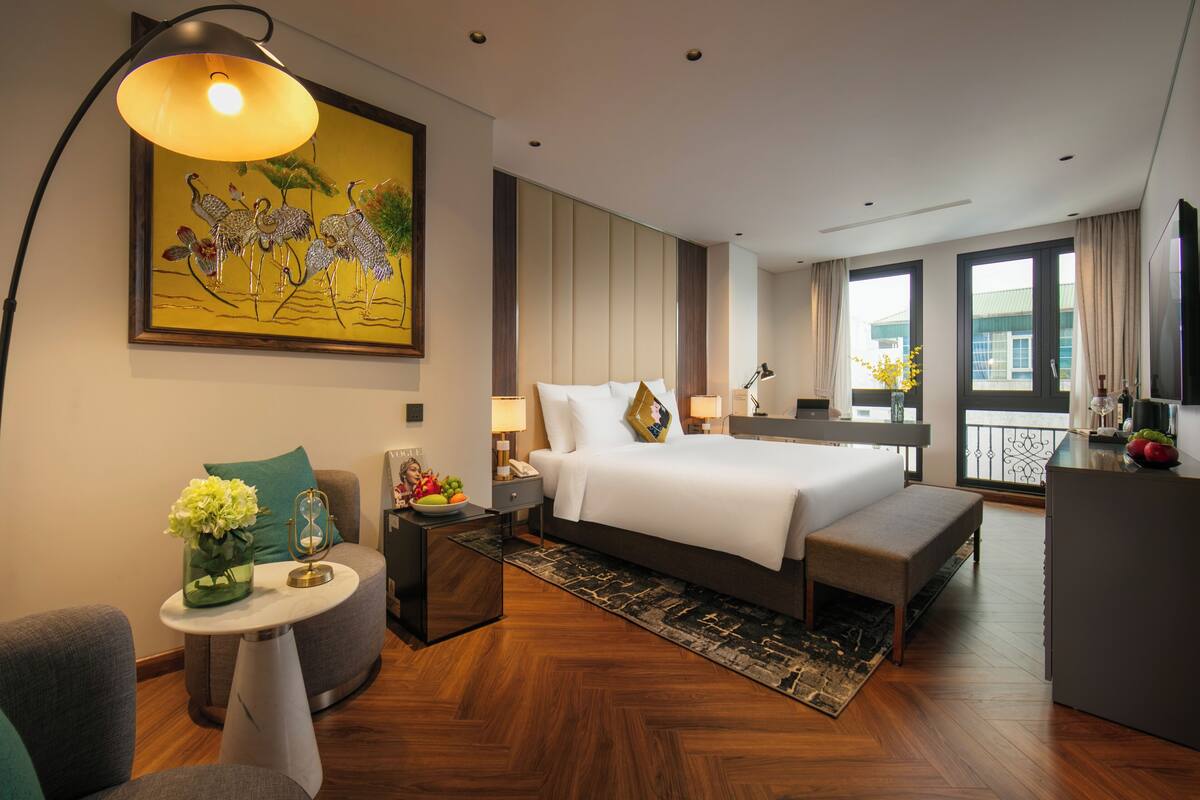 A modern hotel room with a large bed, bedside tables, a small seating area, a painting, and a balcony with a view.