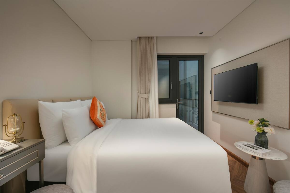 A hotel room with a bed, a desk with a lamp, a TV mounted on the wall, and a small table with a vase of flowers.