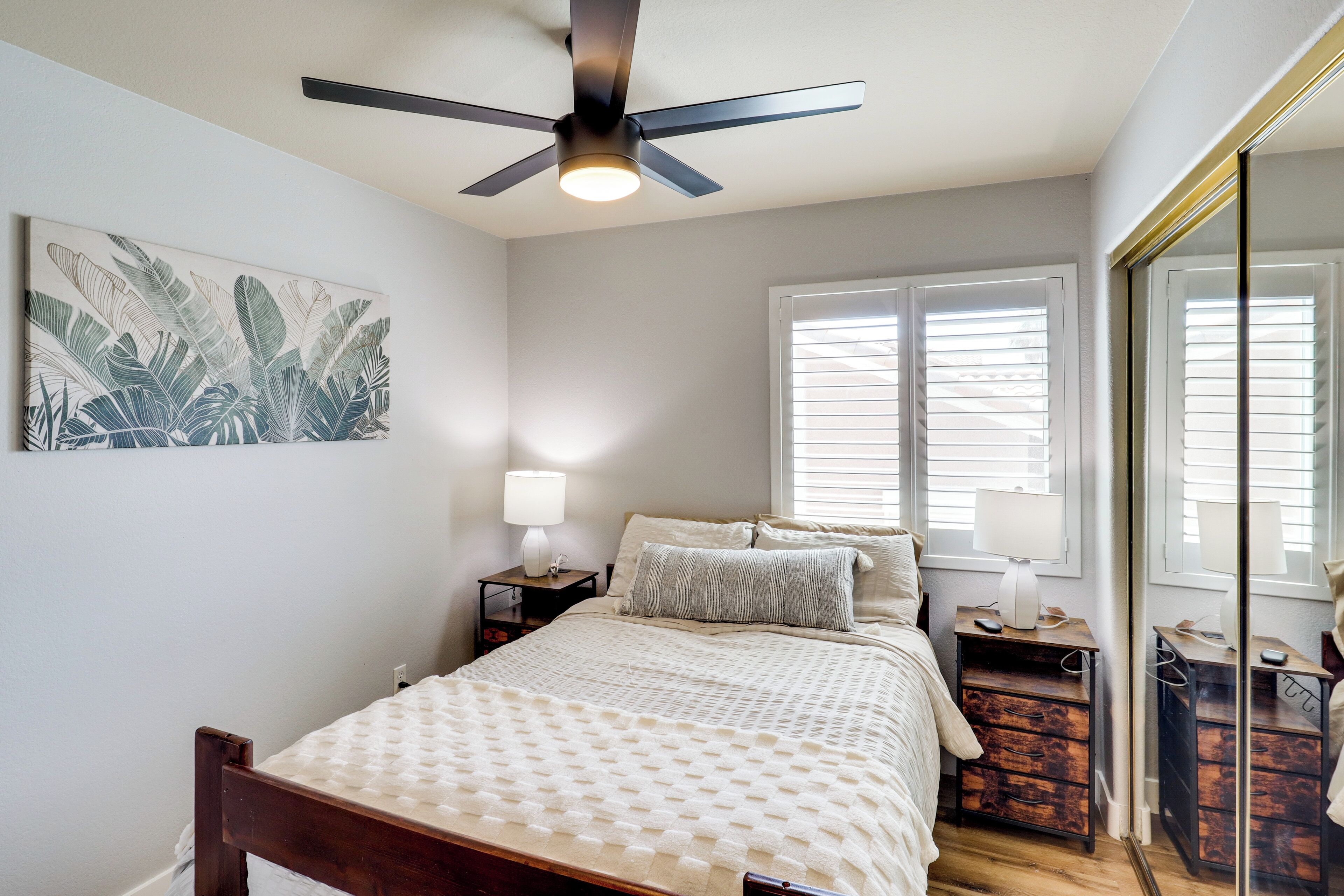 A bedroom with a bed, a ceiling fan, a nightstand, a lamp, a window with blinds, and a mirror.