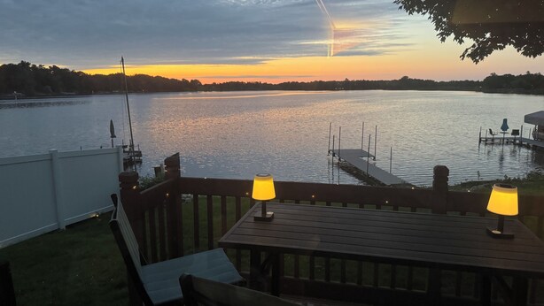 A wooden deck with two table lamps, a dock extending into a calm lake, and a sunset in the background.