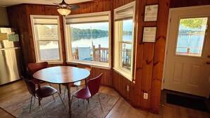 A wooden interior with a dining table and chairs, large windows with a view of a lake, and a refrigerator.