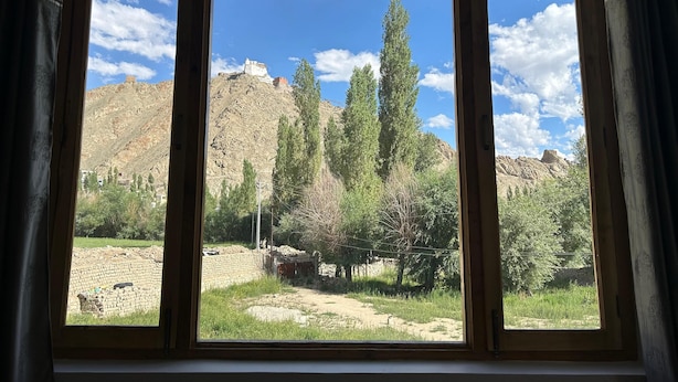 A room with a large window offering a view of a mountainous landscape with trees and a castle-like structure. - Otsal Villa (Leh)