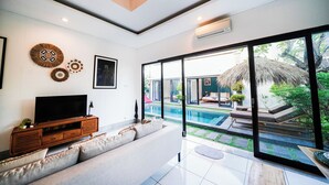 A modern living room with a large TV, a sofa, and a view of a swimming pool and outdoor seating area.