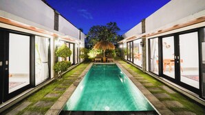 A modern pool area with a clear pool, surrounded by glass doors and a thatched roof structure.