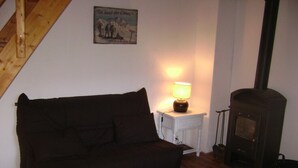 Un salon chaleureux avec un canapé aux couleurs sombres, une petite table d’appoint blanche surmontée d’une lampe, et un poêle à bois.