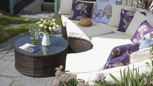 Ein Außenbereich mit Korbmöbeln, einer weißen gepolsterten Couch und violetten floralen Kissen.