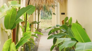 A narrow corridor with a thatched roof, surrounded by lush green plants and a pathway leading to a door. - Lavillas Jogja (Sleman)
