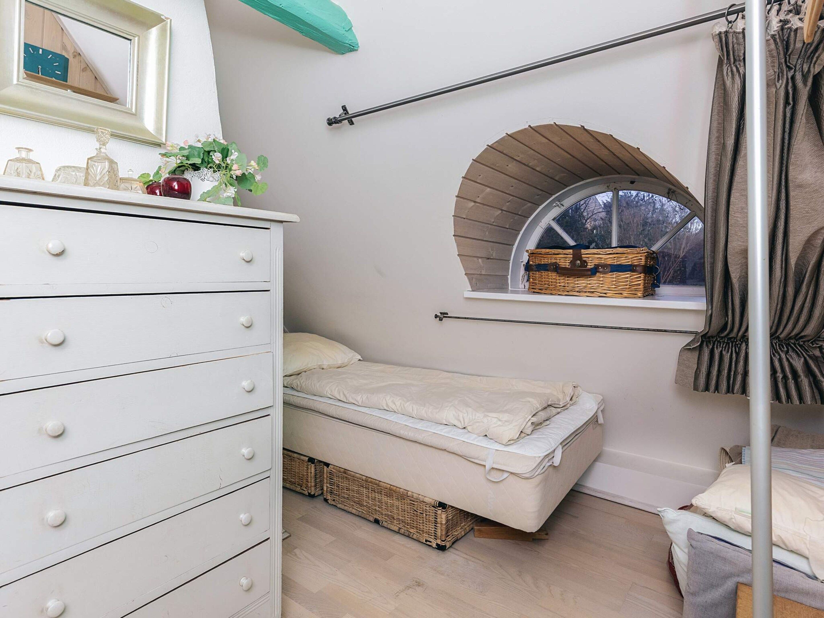 A bedroom with a white dresser, a single bed, a window with a view, and a small arched window above the bed.