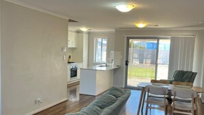 A modern living room with a sofa, a dining table with chairs, a kitchen area, and a sliding glass door leading to an outdoor space. - Secure 4BR w/Garage Whyalla (Whyalla Jenkins)