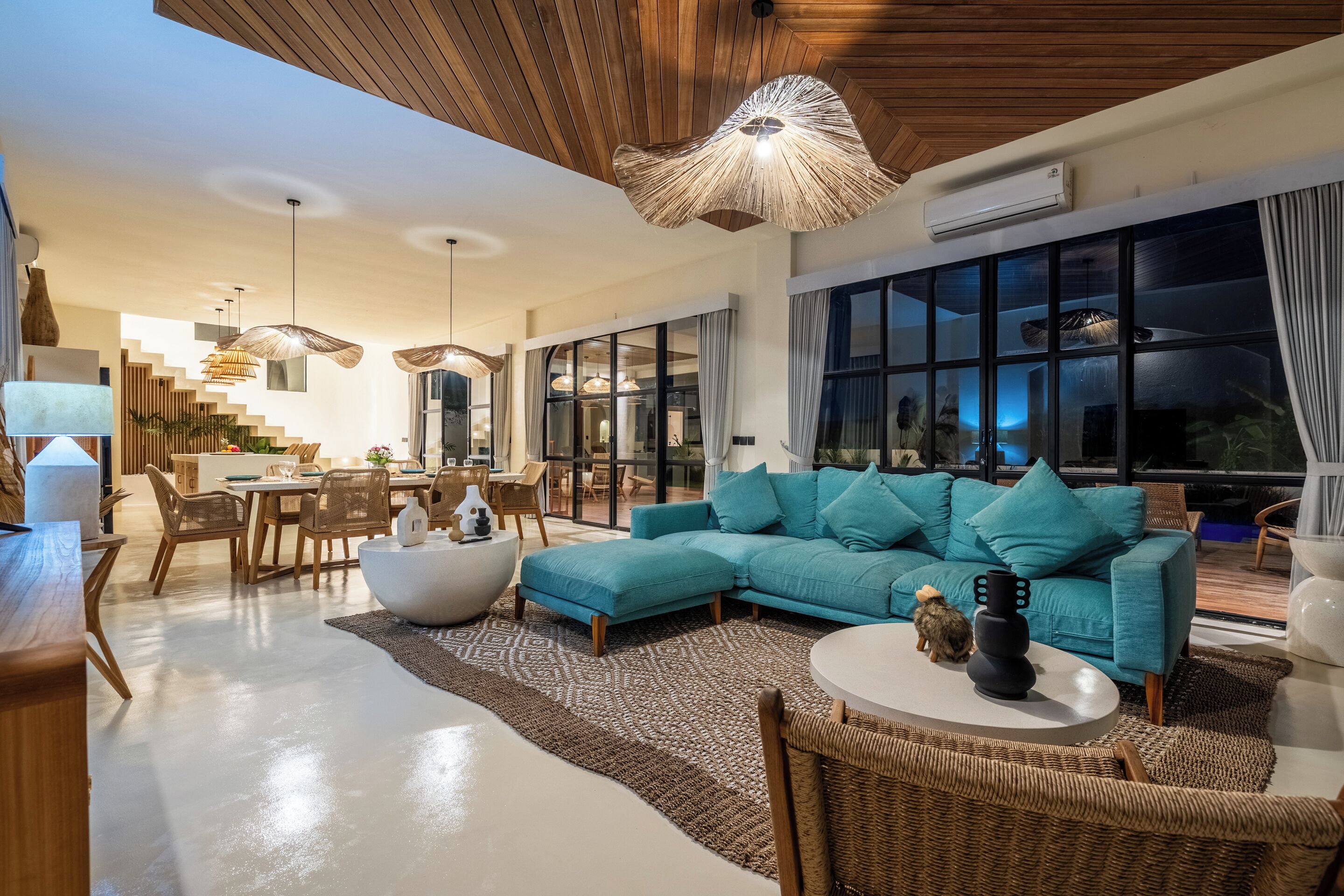 A modern living room with a teal sofa, wicker furniture, and a dining area with a white table and chairs.