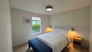 Un dormitorio con cama, mesitas de noche, un banco, una ventana y un aire acondicionado.