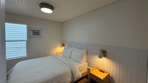Un dormitorio con cama, mesitas de noche, luz empotrada en la pared, ventana con persianas y aire acondicionado.