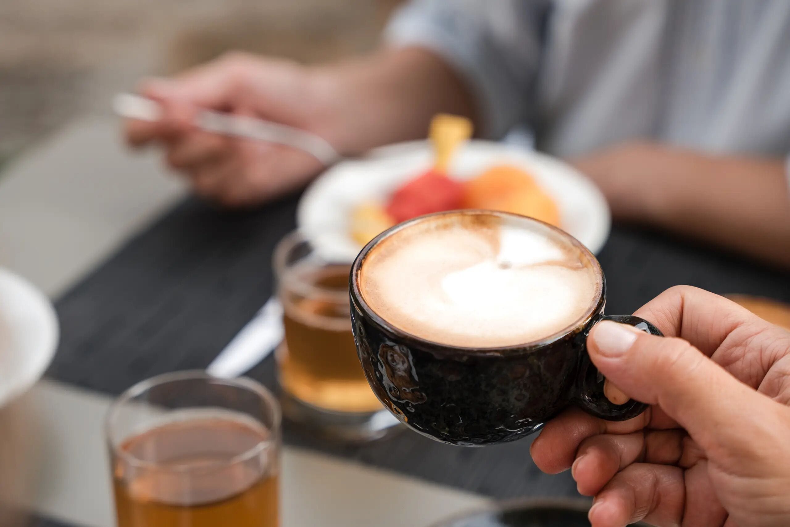 Une personne est assise à une table, tenant une tasse contenant une boisson, une assiette avec de la nourriture et un verre rempli d’un liquide.